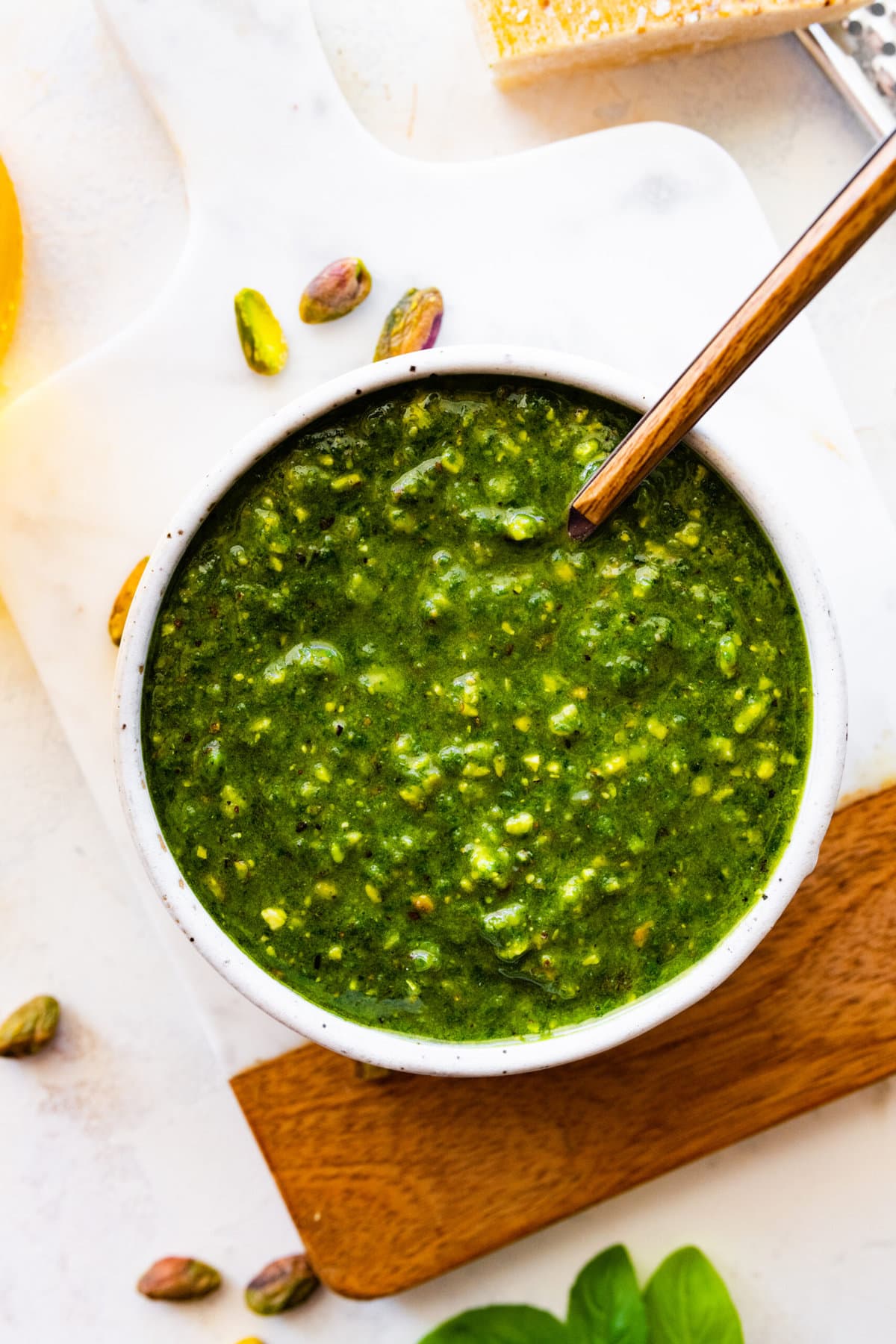 pistachio pesto in a bowl with a spoon in it on a marble and wood cutting board.