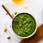 pistachio pesto in a bowl with a spoon in it on a marble and wood cutting board.
