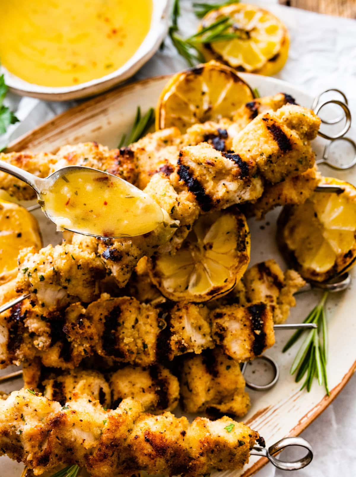 chicken spiedini recipe on a platter with grilled lemons and herbs ready to serve with dipping sauce poured over the top.