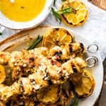 chicken spiedini recipe on a platter with grilled lemons and herbs ready to serve with dipping sauce on the top and dripping off the sides.