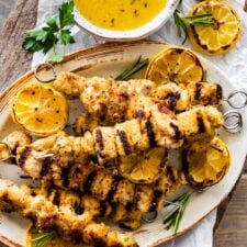 chicken spiedini recipe on a platter with grilled lemons and herbs ready to serve with dipping sauce on the top and dripping off the sides.