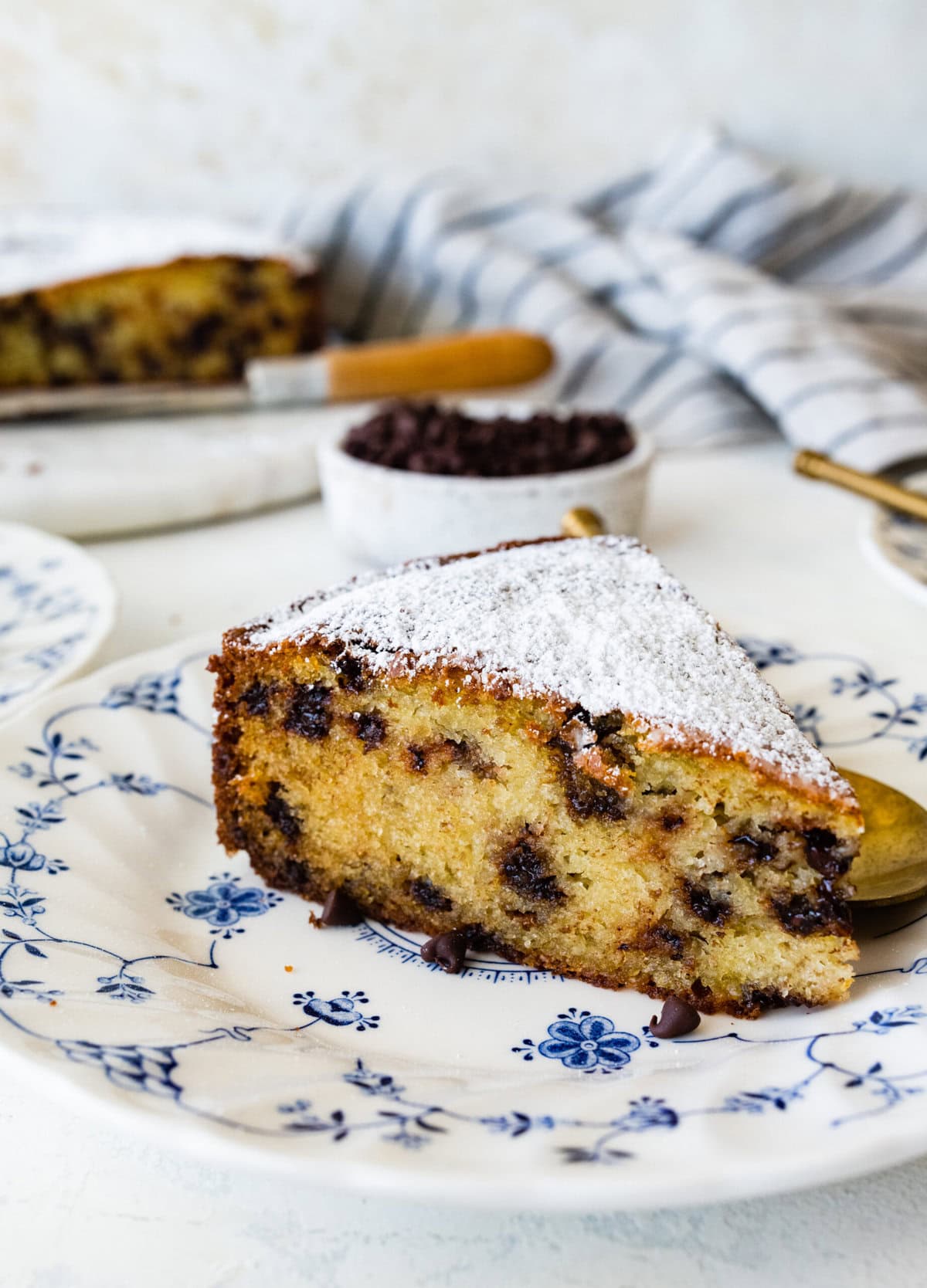 One slice of chocolate chip ricotta cake on a white plate with blue flowers on it. Gold spoon on the plate.