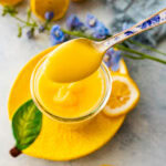 lemon curd in a glass bowl on a lemon shaped plate with a blue spoon in it ready to spoon and serve. Spoon with lemon curd on it taking a scoop.