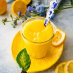 lemon curd in a glass bowl on a lemon shaped plate with a blue spoon in it ready to spoon and serve.
