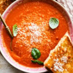 two bowls of creamy tomato soup with basil on top. Spoon in the bowl. Grilled cheese sandwich on the side.
