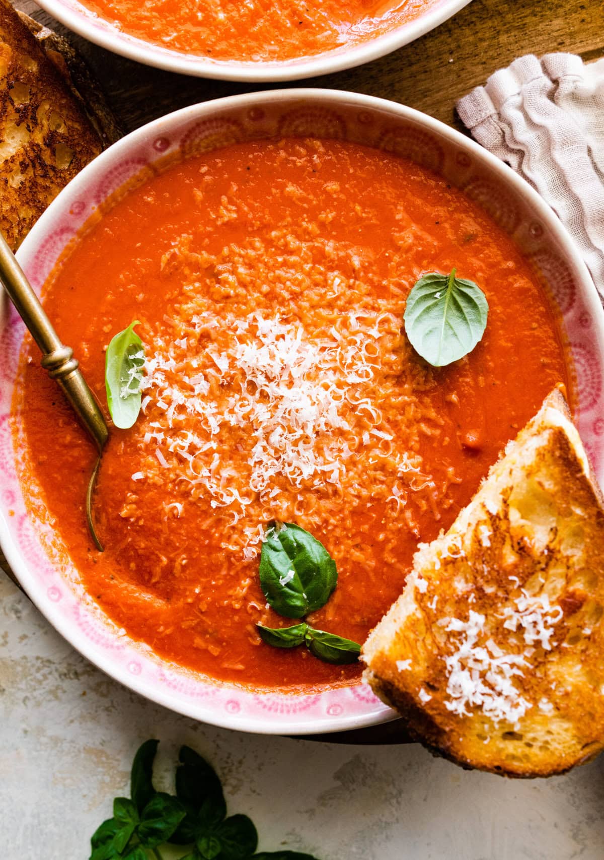 two bowls of creamy tomato soup with basil on top. Spoon in the bowl. Grilled cheese sandwich on the side.