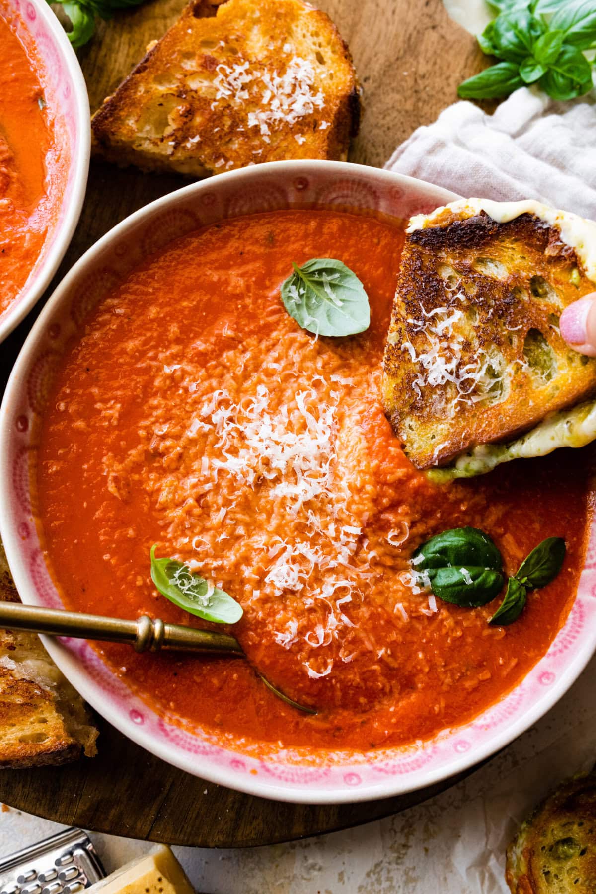 two bowls of creamy tomato soup with basil on top. Spoon in the bowl. Grilled cheese sandwich on the side. Dipping the sandwich in the soup.