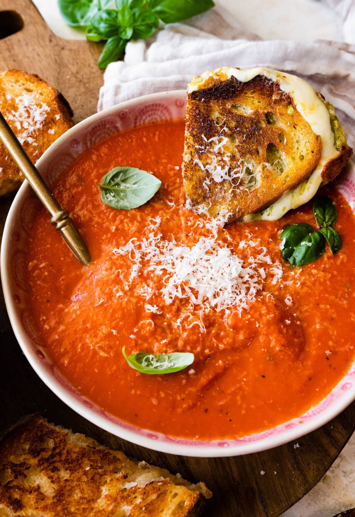 two bowls of creamy tomato soup with basil on top. Spoon in the bowl. Grilled cheese sandwich on the side. Dipping the sandwich in the soup.