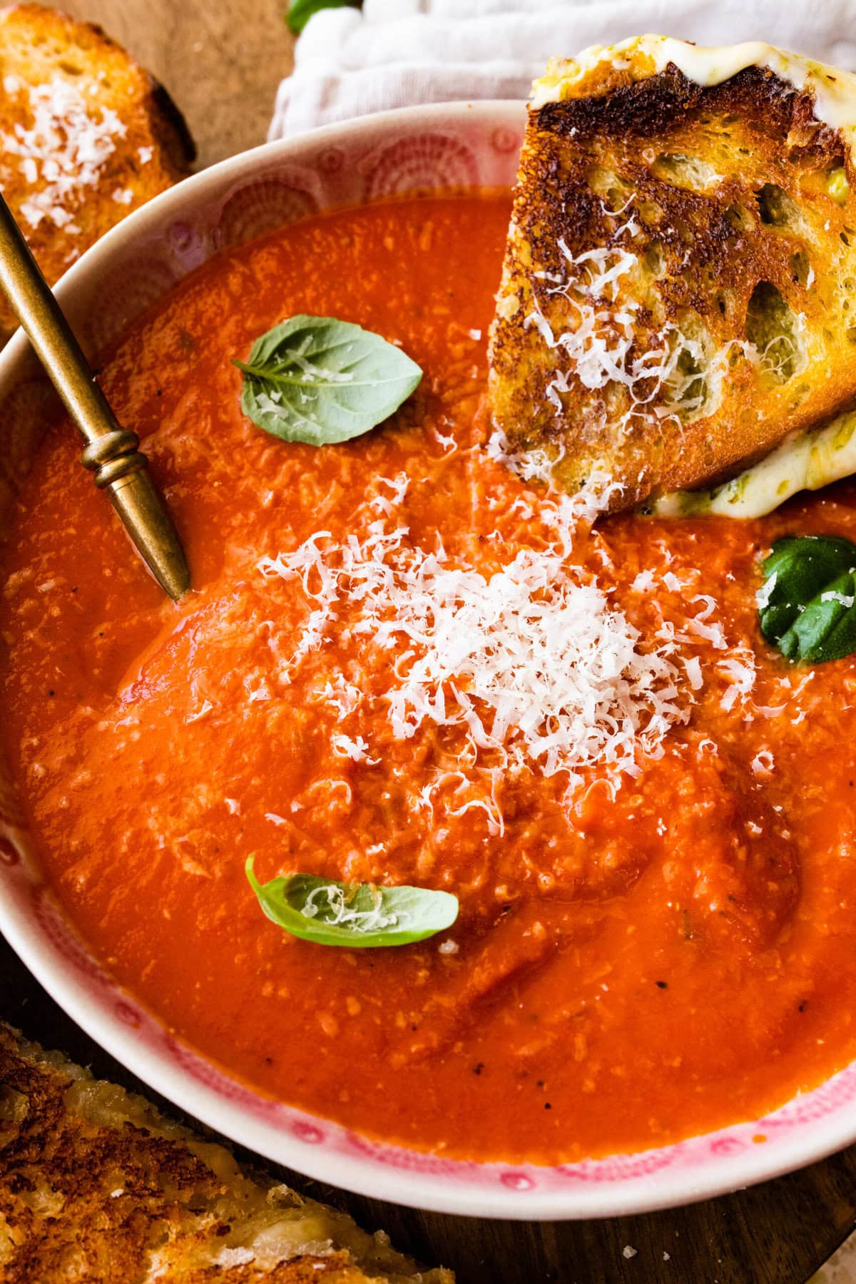 two bowls of creamy tomato soup with basil on top. Spoon in the bowl. Grilled cheese sandwich on the side. Dipping the sandwich in the soup.