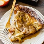 a slice of the apple dutch baby pancake on a white plate with a gold fork ready to eat.