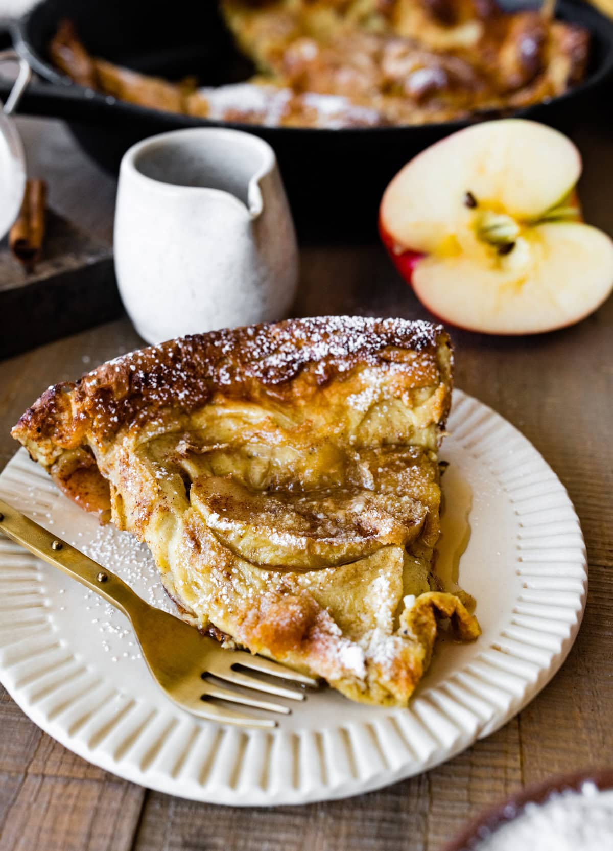 a slice of the apple dutch baby pancake on a white plate with a gold fork ready to eat. Apple cut in half in the background.