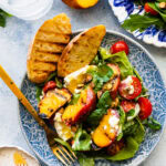plate of grilled peach salad. A small blue and white plate with the salad and burrata. Grilled bread on the side.