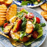 plate of grilled peach salad. A small blue and white plate with the salad and burrata. Grilled bread on the side.