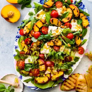 platted grilled peach salad on a blue and white serving platter. Grilled bread and sliced peaches on the side. Salad servers on the side.