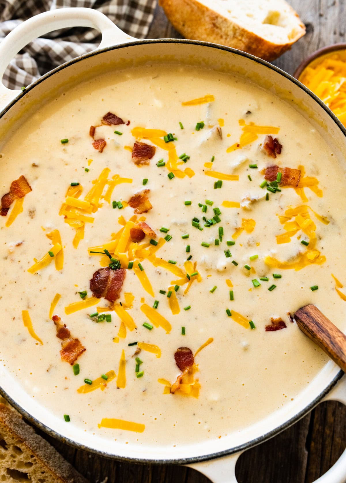 finished baked potato soup with a wooden spoon in it. Cheddar cheese and bacon bits on top. Bread on the side.