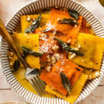 plated pumpkin Ravioli with Sage Brown Butter Sauce Recipe on a white plate. Served with parmigiano cheese on top. Fork in the plate ready to eat. Extra hazelnuts and sage on the side.