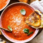 two bowls of creamy tomato soup with basil on top. Spoon in the bowl. Grilled cheese sandwich on the side.