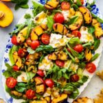 platted grilled peach salad on a blue and white serving platter. Grilled bread and sliced peaches on the side. Salad servers on the side.