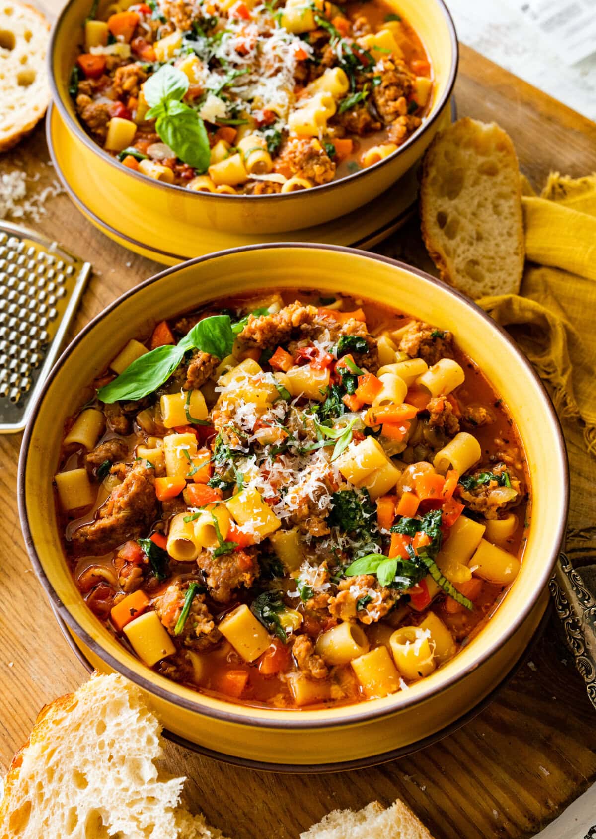 A bowl of the Italian sausage soup ready to eat with a leaf of basil on the side. Parmigiano cheese and bread on the side for serving.