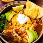 Easy Mexican chicken soup in a bowl with all the toppings on it and a spoon in it ready to eat. Avocado, cheese, sour cream and chips in the bowl.