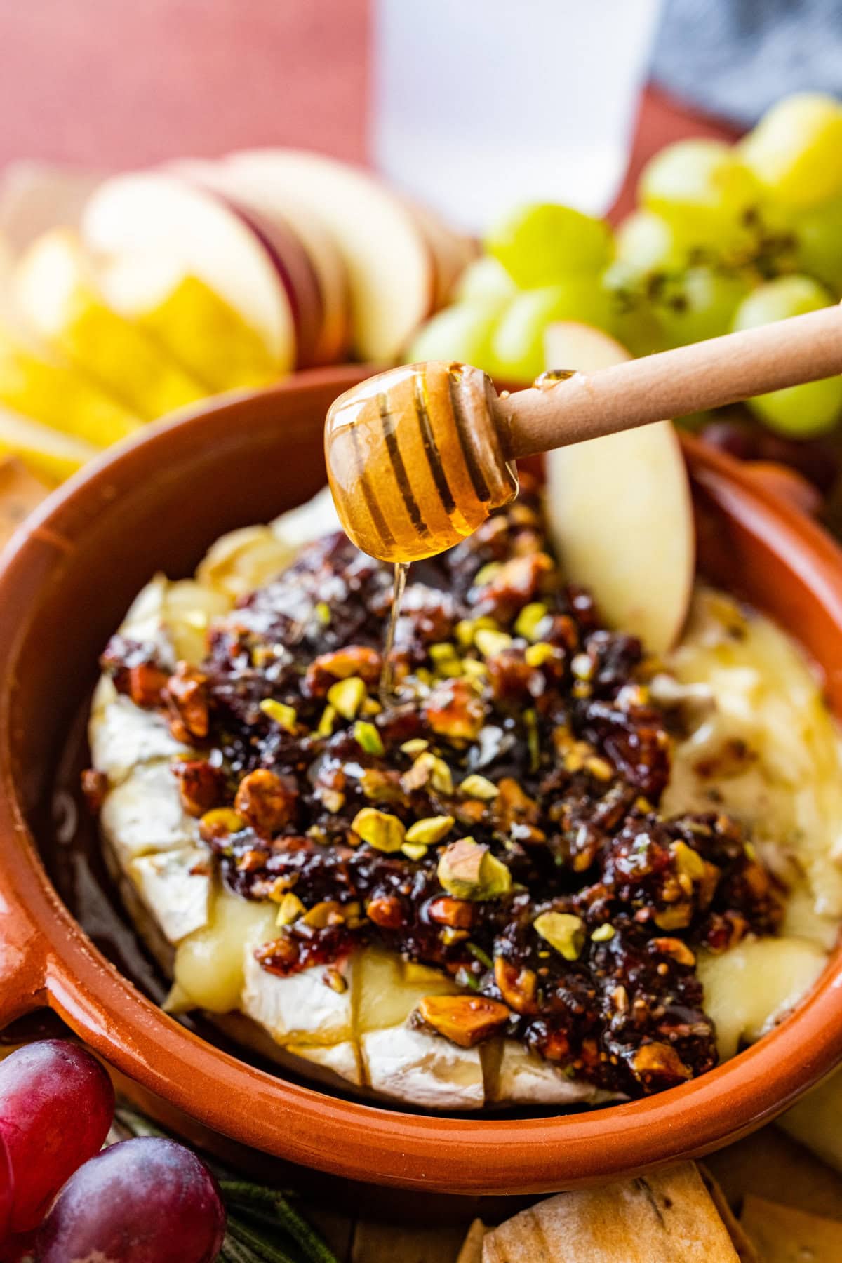 Honey drizzled on top of the beautiful baked brie with the topping on a platter with fruits, crackers, and a cheese knife to serve.