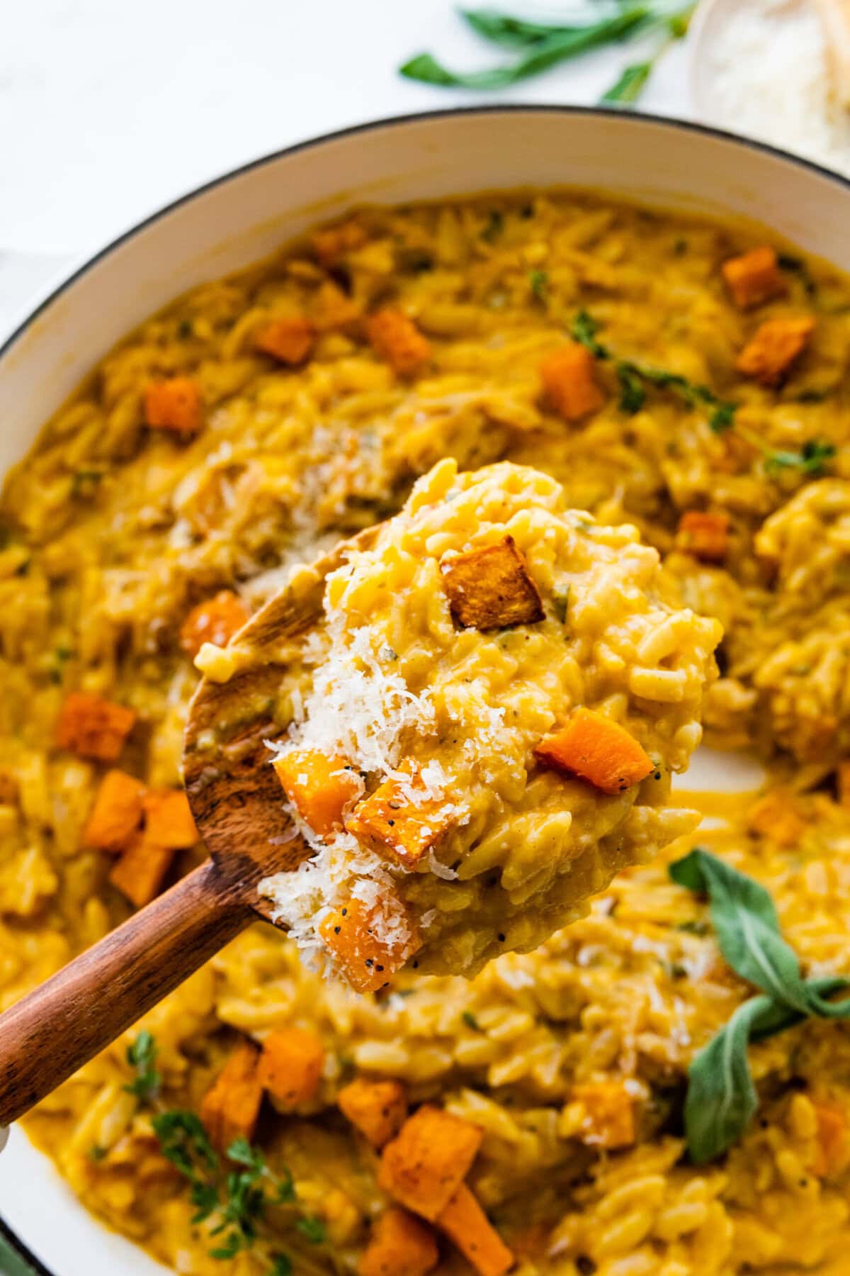 finished dish in the pot with sage on top. Wooden serving spoon in the pot. Spoon holding a portion of the orzo dish ready to serve.