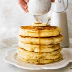 a big stack of fluffy pancakes on a plate with butter on top. Pouring hot syrup on the top. A jug of milk visible in the background.
