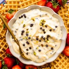 Spoon in a bowl of cannoli dip on a platter with strawberries and pizzelle cookies as dippers.