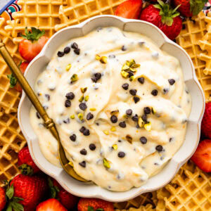 Spoon in a bowl of cannoli dip on a platter with strawberries and pizzelle cookies as dippers.