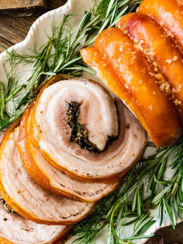 Beautiful porchetta roast with crispy skin and juicy interior and herb layer on a platter ready to serve. Fresh herbs around the white platter.