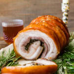 Beautiful porchetta roast with crispy skin and juicy interior and herb layer on a platter ready to serve. Fresh herbs around the white platter.