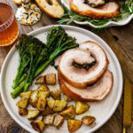 Beautiful porchetta roast with crispy skin and juicy interior and herb layer on a platter ready to serve. Fresh herbs around the white platter.