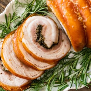Beautiful porchetta roast with crispy skin and juicy interior and herb layer on a platter ready to serve. Fresh herbs around the white platter.