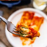 Baked Spinach and ricotta manicotti on a plate with a fork taking a bite. Fork taking the bite. Fresh parmigiano cheese on the side.