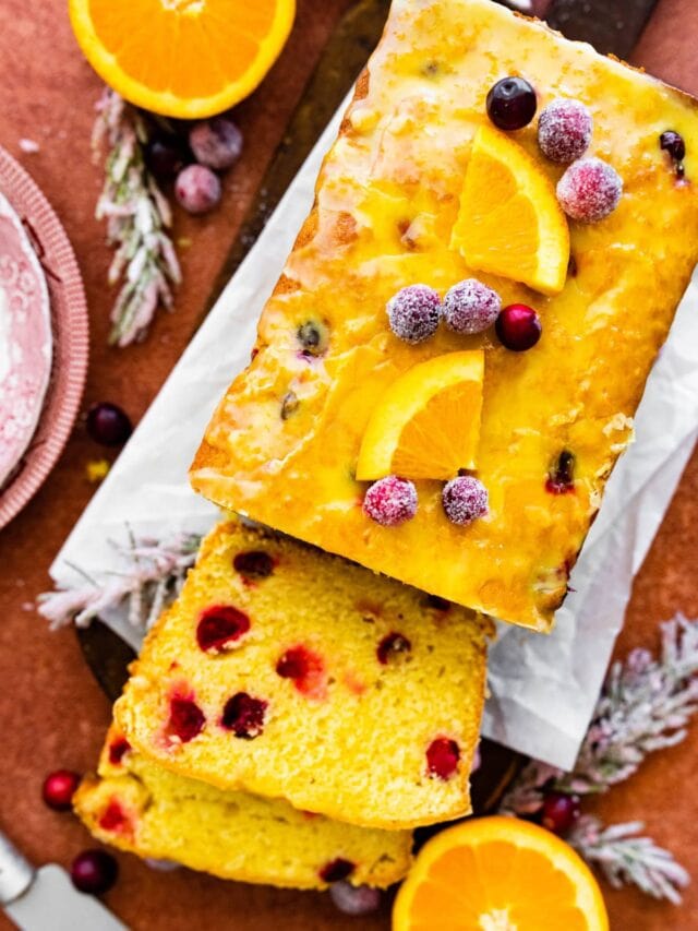 beautiful cranberry orange bread with orange glaze on a terracotta colored background. Oranges and sugared cranberries as decoration. A slice taken out of the bread.