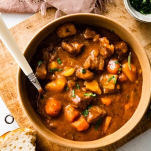 A big bowl of beef stew with a little fresh parsley on top. Bread on the side. A spoon in the bowl ready to eat.