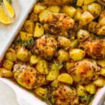 Baked Chicken and potatoes recipe in a white baking dish. The potatoes and chicken have a golden brown top with herbs as a garnish. A side of lemons for serving.