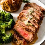 Sliced New York steak on a plate with butter sauce on top. There is a side of broccoli and baked potato on the side.