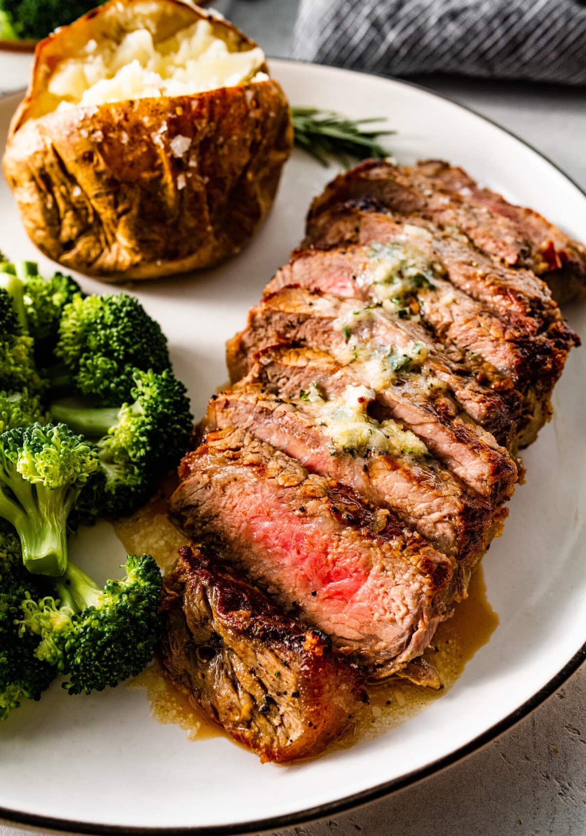 Sliced New York steak on a plate with butter sauce on top. There is a side of broccoli and baked potato on the side.