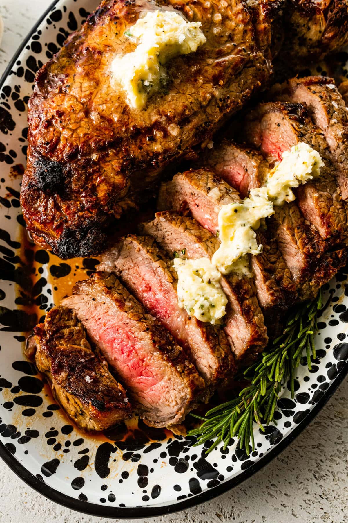 Sliced broiled New York Steak on a platter. One steak is cut into slices and there is a butter sauce pored over the top.