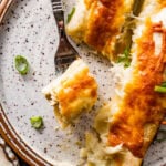 White chicken enchilada on a plate with a fork. Limes on the side and ready to eat. A bite taken out of the enchiladas with a fork.