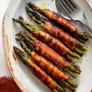 Bacon wrapped asparagus on a serving platter with serving utensils. Crispy brown edges and sauce.