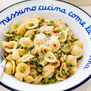 a blue and white Italian style plate with a portion of the creamy pasta with pancetta and peas. Extra cheese and red pepper flakes on top. Close up of the plate with the pasta.