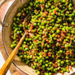 peas and pancetta recipe in a large white serving bowl with a wooden spoon ready to serve. Serving bowl on a wooden board.
