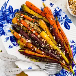 Roasted rainbow carrots after cooking in the oven on a beautiful white and blue floral serving platter. Carrots are roasted perfectly. Add hazelnuts on top for crunch. Serving utensils on the side.