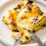 slice of white chicken lasagna on a plate with a fork and knife that cut one piece to eat.