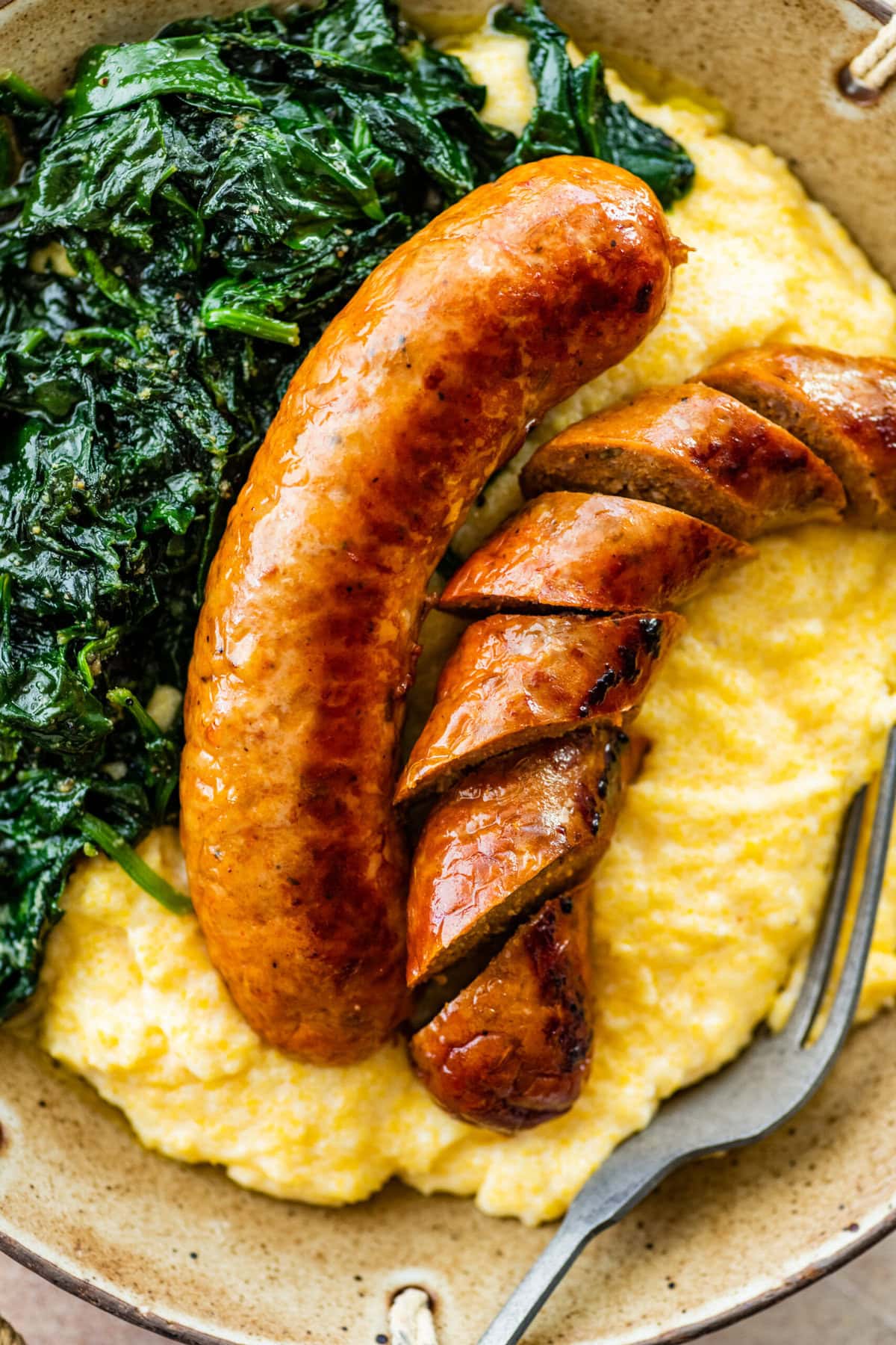 Italian baked sausage in a bowl with polenta and cooked greens. Sausage is cut into slices.