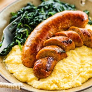 Italian baked sausage in a bowl with polenta and cooked greens. Sausage is cut into slices.