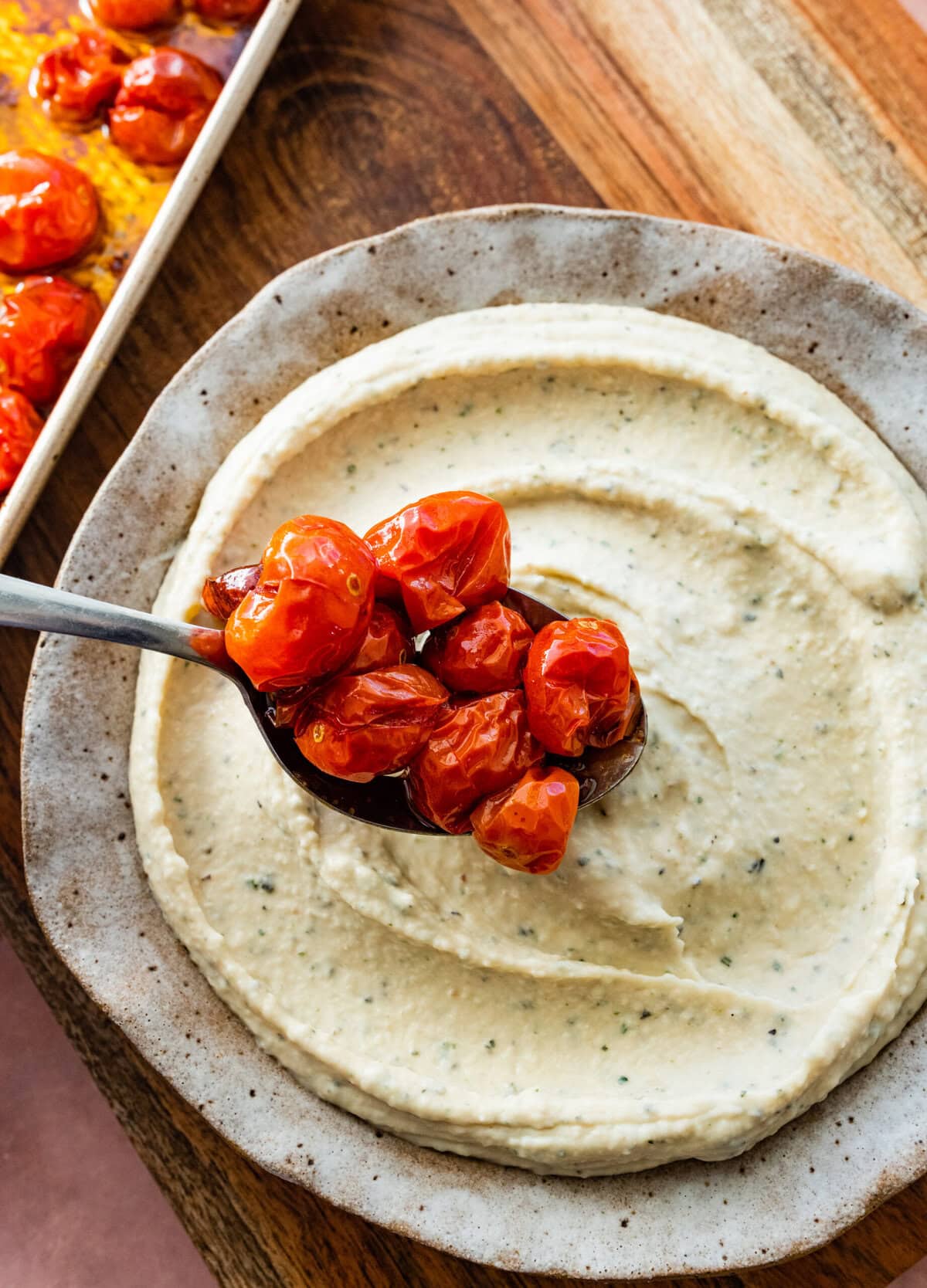 white bean dip recipe on a platter with spoon placing the blistered tomatoes on top.
