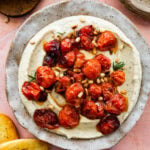 creamy white bean dip on a serving plate with the blistered roasted tomatoes on top on a pink background. Crostini on the side.